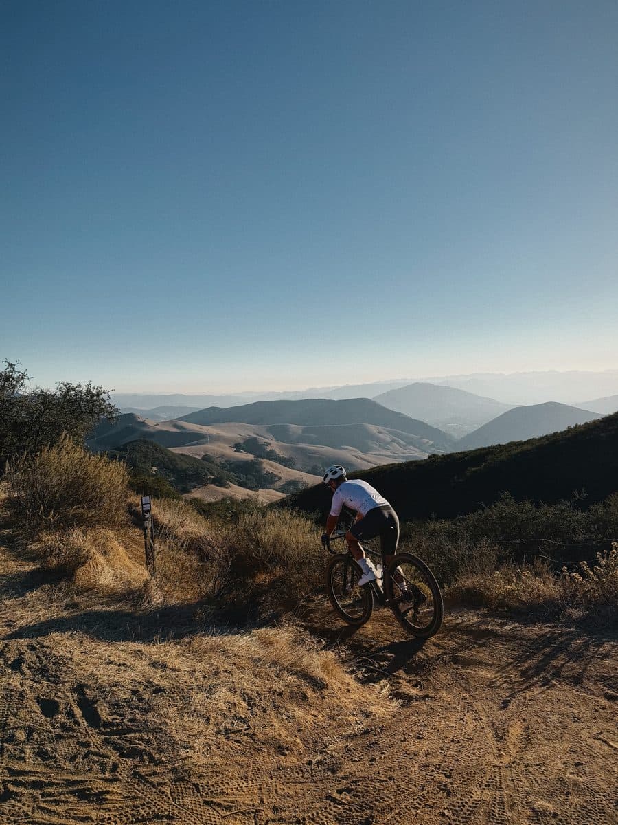 A gravel rider, seen from behind, dropping onto a dirt trail above the hills